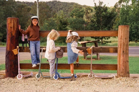 Three children line up against a wooden farm-style fence with their Banwood aluminium scooters in a range of soft pastel colours