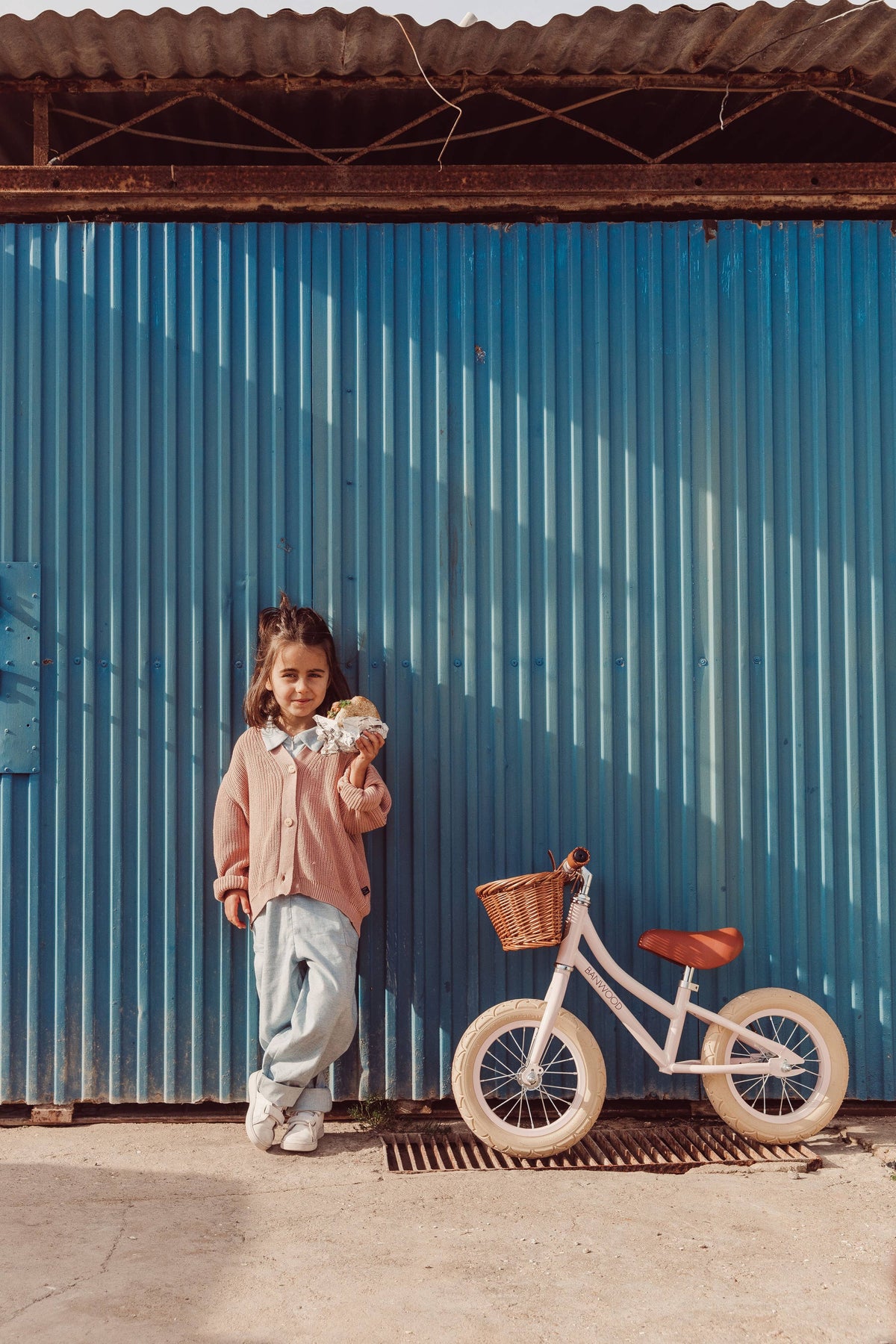 Child standing next to a scandi chic  pale pinkbalance bike  from Banwood with a wicker basket and leather saddle against a blue corrugated metal wall.. 